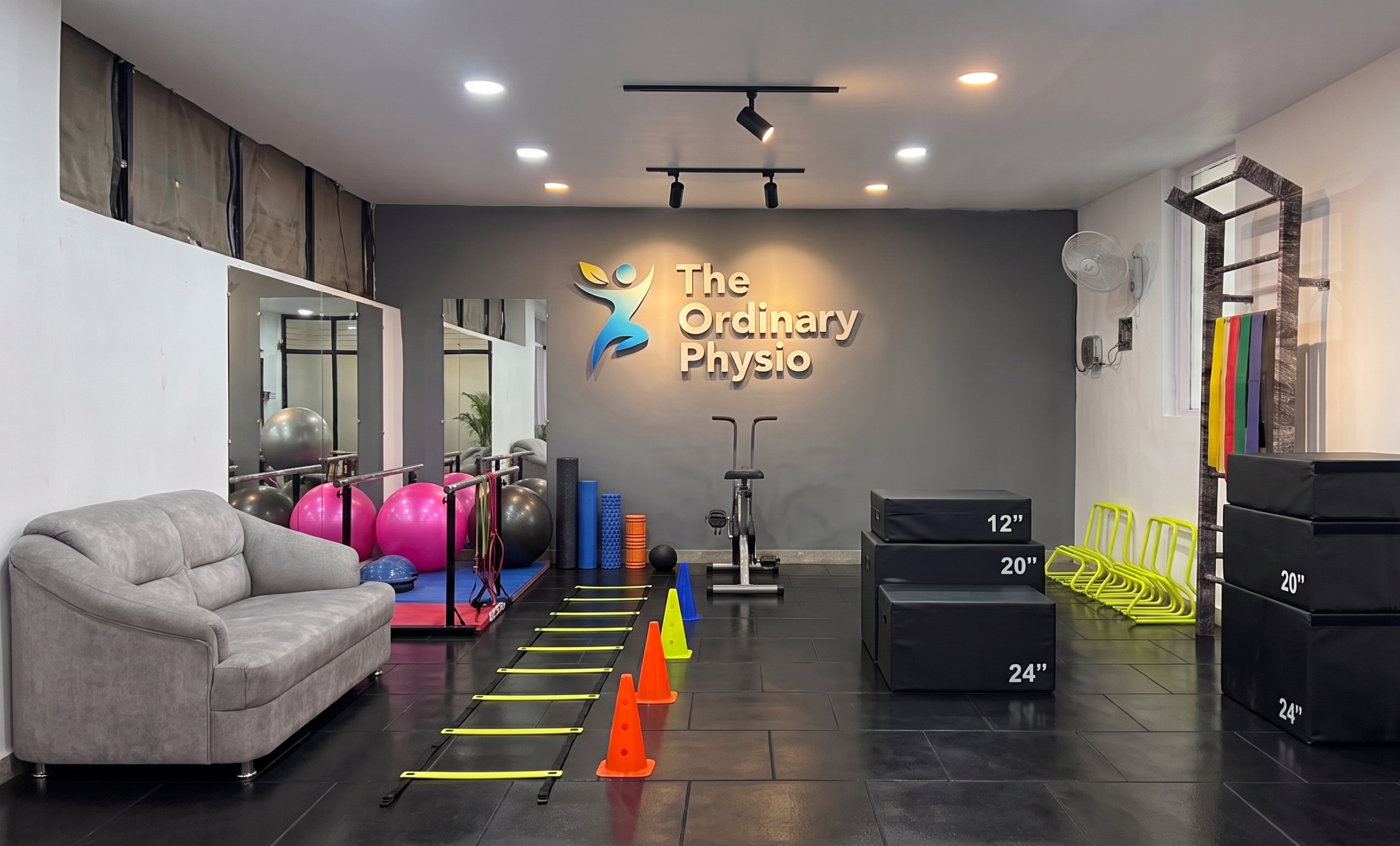 Wide-angle photograph of the modern, well-equipped physical therapy studio, 'The Ordinary Physio'. The foreground shows an agility ladder with orange and yellow cones. The left side has a grey sofa and mirrors reflecting stability balls (pink and grey), medicine balls, and foam rollers on a blue mat. The center-back wall displays the gold text 'The Ordinary Physio' with a stylized icon above a stationary exercise bike. To the right is a wall-mounted ladder with resistance bands and a stack of black soft-sided plyometric boxes. Option 2 (Focused on Core Services): Modern rehabilitation and physiotherapy studio 'The Ordinary Physio' interior view, featuring extensive exercise equipment including stability balls, foam rollers, an exercise bike, stacked soft plyometric boxes, an agility ladder, and resistance bands.